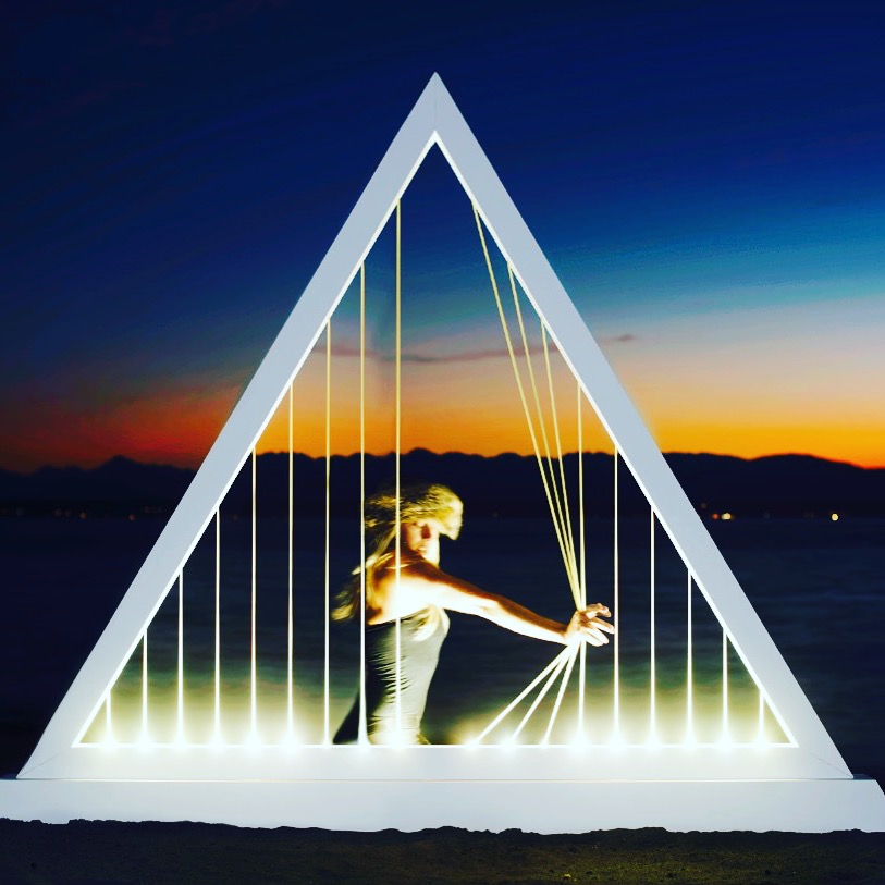 Jasmine Zimmerman performing inside a glowing triangular Elastic Light installation at the water's edge at dusk